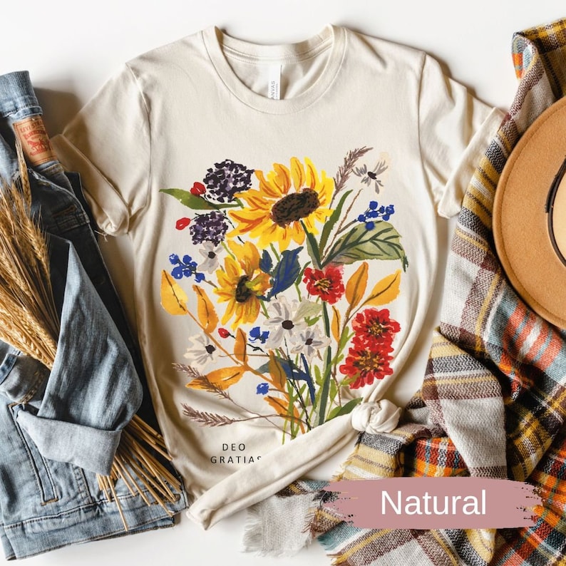 A women's natural colored t-shirt featuring a loose watercolor floral bouquet with yellow sunflowers and berries. The words "Deo Gratias" are at the bottom of the t-shirt. Shirt is styled with a jean jacket, a brown wool hat and plaid scarf For Fall