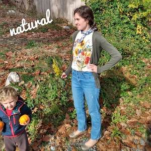 Puede incluir: Una mujer lleva una camisa blanca con estampado floral, una chaqueta de punto verde y unos vaqueros azules. Est&aacute; de pie en un jard&iacute;n con hojas ca&iacute;das y plantas verdes. La palabra "natural" est&aacute; escrita en blanco sobre un fondo marr&oacute;n en la esquina superior izquierda de la imagen. Un ni&ntilde;o peque&ntilde;o est&aacute; de pie en la esquina inferior izquierda de la imagen, sosteniendo dos peque&ntilde;as calabazas naranjas.