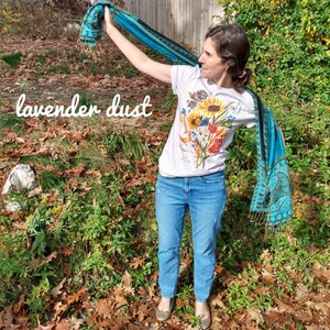 Puede incluir: Una mujer lleva una camiseta blanca con un estampado floral y el texto "Old Flowers" y sostiene una bufanda azul y negra. La bufanda est&aacute; adornada con dise&ntilde;os de paisley. La mujer est&aacute; de pie en una zona cubierta de hierba con hojas ca&iacute;das.