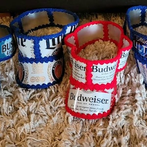 May include: Seven crocheted visors made from beer can tabs. The visors are in red, blue, and white. The visors feature various beer brands, including Budweiser, Busch Light, Coors Light, and Natural Light.