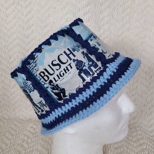 May include: A blue and white crocheted hat with a Busch Light beer can label. The label features a winter scene with the text "Busch Light" and "Snow Day".