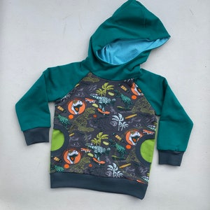 Könnte beinhalten: Ein türkisfarbener Kapuzenpullover mit einem grauen und grünen Dinosaurier-Druck. Der Druck zeigt Dinosaurier beim Skateboardfahren und Palmen. Der Pullover hat ein zweifarbiges Design in Grün und Grau.