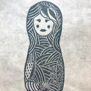 Puede incluir: Grabado en linóleo en blanco y negro de una muñeca rusa con un diseño de conchas marinas. La muñeca está rodeada de conchas marinas y algas marinas. La impresión está firmada "Sister Sea" y "110 L. Oyen".