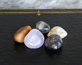 May include: A collection of six polished gemstones in various colors, including brown, purple, green, and black. The stones are arranged on a black leather-bound book with gold trim. The background is a soft, textured, light gray.