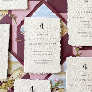 May include: A collection of wedding invitations with textured white paper cards, embossed with floral patterns and the names ILARIA PELLEGRINI and GIACOMO CORDONI. The cards are arranged with burgundy envelopes and floral decorations, with a watercolour landscape.