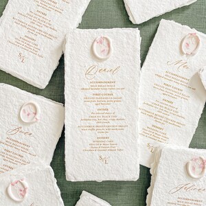 May include: A collection of elegant wedding menus on textured white paper. Each menu features gold text, a floral wax seal, and a detailed menu. The menus are arranged on a green surface, creating a sophisticated aesthetic.