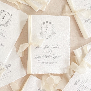 May include: Ivory-colored wedding programs with elegant calligraphy and a floral crest. Each program is tied with a delicate ribbon. The programs feature the names "Riazi's Aidile Barker and Logan Stephen Taplett" and the date "December 27th, 2023".