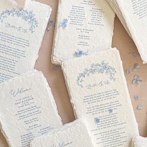 May include: A collection of rectangular wedding programs printed on off-white handmade paper with deckled edges. The text is in dark blue ink, featuring floral illustrations and the title Build My Life. Scattered light blue hydrangea petals are visible.