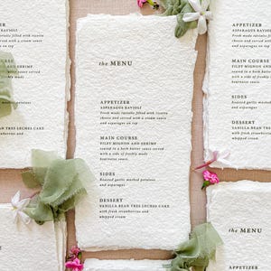 Peut inclure: Menus en papier faits à la main avec des bords déchirés, présentant les détails du menu en texte noir élégant. Les cartes sont ornées de tissu vert et de petites fleurs roses et blanches, créant une esthétique rustique et élégante. La marque "KATTONE PAPER" est visible.