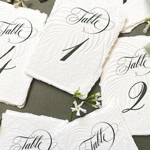May include: White textured paper table numbers with elegant calligraphy. Each card features a unique number in dark green, ranging from 1 to 11. The word "Table" is written in cursive above the numbers. The cards are arranged on a green surface.