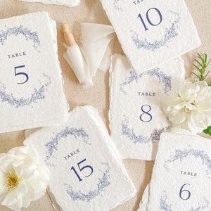 Peut inclure: Cartes de numéros de table en papier blanc avec des motifs de couronnes florales bleues et des numéros de table. Les numéros vont de 5 à 15. Un objet en bois et des fleurs blanches sont également visibles.