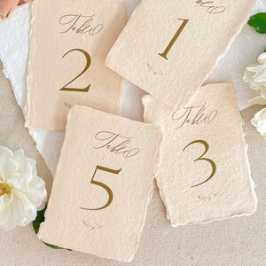May include: Five table number cards with a rustic, cream-colored background and gold lettering. The cards are numbered 1, 2, 3, and 5. Each card has the word "Table" written in cursive above the number.