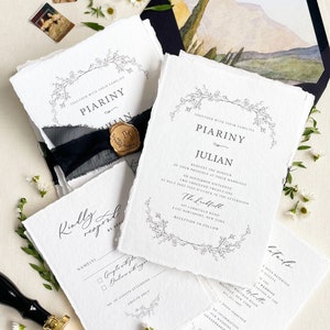 May include: A wedding invitation suite with white, textured paper and black calligraphy. The set includes invitation, RSVP, and details cards, tied with black ribbon and sealed with a gold wax seal. The envelope features a watercolour landscape scene.