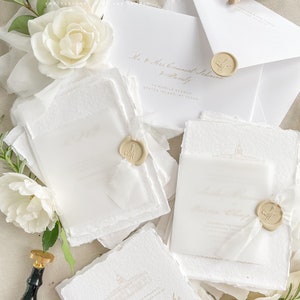 May include: A stack of white wedding invitations with gold lettering and wax seals. The invitations are on textured paper and have a delicate, elegant design. The text on the invitations reads "The honor of your presence is requested at the marriage of Sasha Hernandez and Lauren Chapparelli".
