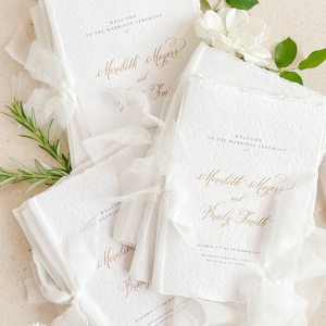 May include: White wedding ceremony programs with gold calligraphy text. The programs are folded and tied with a white ribbon. The text reads "Welcome to the marriage ceremony of Meredith Magers and Brady Smith".