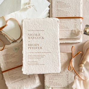 Luxury colored handmade paper wedding invitation suite styled in an editorial flat lay featuring textured cotton deckle edge invitations with warm terracotta typography, paired with details and RSVP cards, ivory envelopes and silk ribbon accents on neutral linen background.