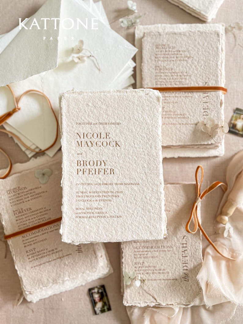 Luxury colored handmade paper wedding invitation suite styled in an editorial flat lay featuring textured cotton deckle edge invitations with warm terracotta typography, paired with details and RSVP cards, ivory envelopes and silk ribbon accents on neutral linen background.
