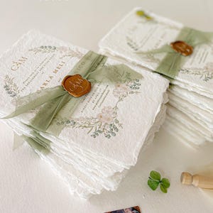 May include: A stack of white, textured paper invitations with a green ribbon and a wax seal. The invitations feature a floral design with a watercolour effect. The text on the invitation reads "C&M" and "Carla Meli and Nico".