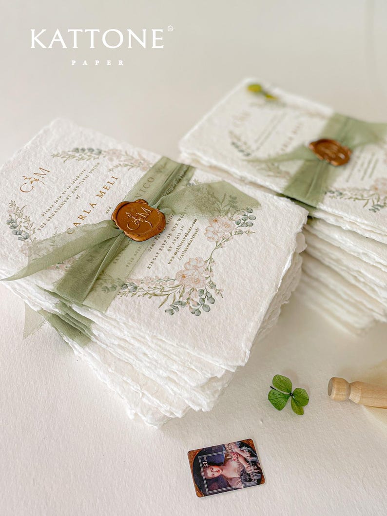 May include: A stack of white, textured paper invitations with a green ribbon and a wax seal. The invitations feature a floral design with a watercolour effect. The text on the invitation reads "C&M" and "Carla Meli and Nico".