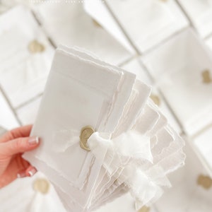 May include: A stack of white, textured paper cards with a gold wax seal and a white ribbon. The cards are arranged in a fan shape and have a slightly distressed look. The text "PARADISE INVITATIONS FINE ART STUDIO" is visible at the top of the image.