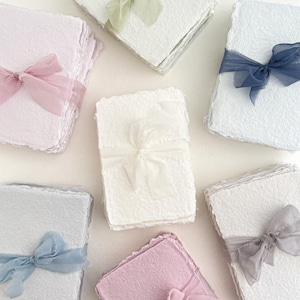May include: Handmade paper stacks, each bound with a sheer ribbon. The paper is in pastel shades of pink, blue, and white. Ribbons in matching colours of pink, blue, green, and grey adorn each stack, creating a soft, elegant aesthetic.