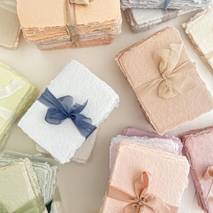 May include: A collection of pastel-coloured handmade paper stacks, each bound with a ribbon. The paper features a textured finish and a deckled edge. Ribbons are in shades that complement the paper colours, such as peach, pink, and blue. The overall look is delicate.