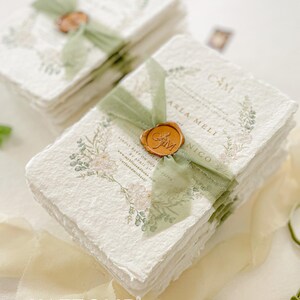 May include: Stack of white paper invitations with green ribbon and a wax seal. The invitations have a floral design and the text "Camille and Carla Meli" is visible. The invitations are on a white surface with a green ribbon.