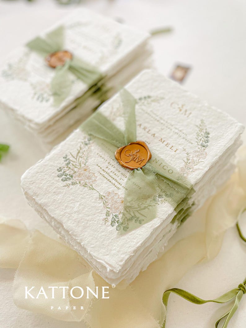 May include: Stack of white paper invitations with green ribbon and a wax seal. The invitations have a floral design and the text "Camille and Carla Meli" is visible. The invitations are on a white surface with a green ribbon.