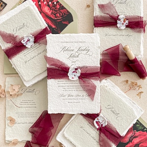 May include: Wedding invitation suite featuring handmade paper with deckled edges. Each invitation is tied with a burgundy ribbon and sealed with a wax stamp. The suite includes details cards and an envelope with a floral design.