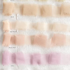 May include: Close-up of several rows of ruffled fabric ribbon in pastel shades: oyster, mink, soft peach, dusty pink, lilac, and white. The fabric has a delicate, sheer appearance with frayed edges.