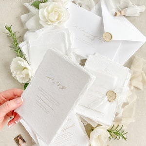 May include: White wedding invitation suite with gold calligraphy and wax seals. The invitation includes the couple's names, wedding date, and RSVP information. The invitation is on textured paper with a rustic, handmade look.