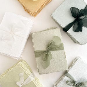 May include: Handmade paper stacks in pastel shades, each bound with a coordinating ribbon. The paper has a textured, organic appearance, with colours including white, yellow, green, and blue. The ribbons add a delicate touch to the stationery.