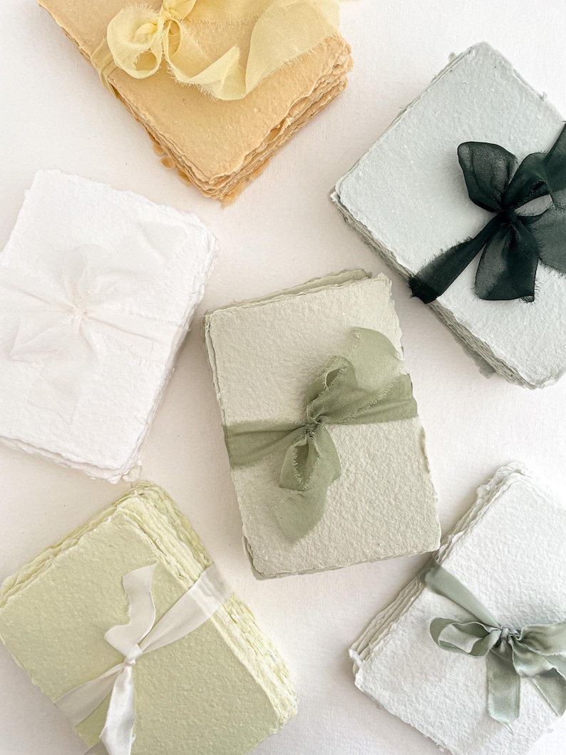 May include: Handmade paper stacks in pastel shades, each bound with a coordinating ribbon. The paper has a textured, organic appearance, with colours including white, yellow, green, and blue. The ribbons add a delicate touch to the stationery.