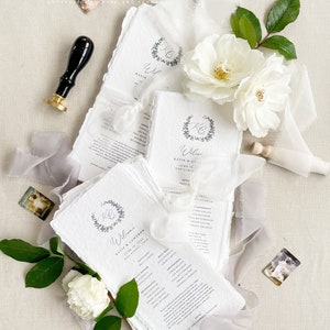 May include: Wedding ceremony program cards with a white background and gray text. The cards feature a floral wreath with the initials "K & C" and the words "Welcome Katie & Cameron June 11, 2023 New Albany, Ohio".