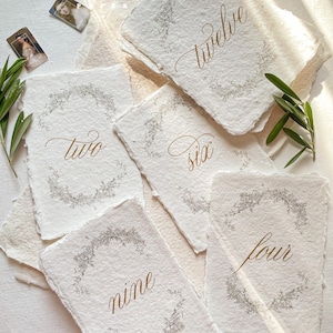 May include: Handmade paper table numbers with gold calligraphy. Each card features a floral wreath design around the number. Numbers visible are two, four, six, nine, and twelve. The paper has a textured, deckled edge. The brand name "KATTONE" is visible in the top left corner.