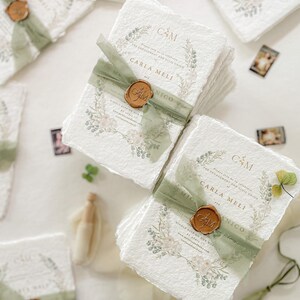 May include: A stack of white, textured paper invitations with a green ribbon and a wax seal. The invitations feature a floral design and the text "Carla Meli & Nico".