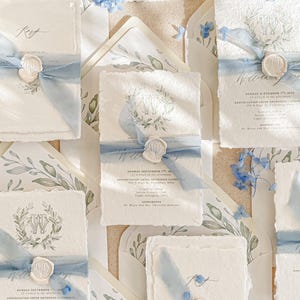 May include: Elegant wedding invitations with a watercolor floral design, featuring a light blue ribbon and wax seal. The invitations have a textured, off-white paper with the text "KATTONE PAPER" at the top. The overall theme is a classic, romantic wedding.