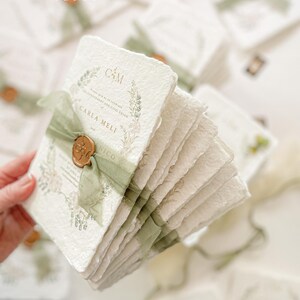 May include: A stack of white, textured paper invitations with green ribbon and a gold wax seal. The invitations have a floral design and the text "C&M" and "Carla Meli" printed on them.