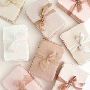 May include: A collection of handmade paper stacks in cream, pink, and peach hues, each bound with silk ribbons. The paper features a textured, deckled edge. Ribbons in matching tones enhance the soft, elegant look. Suitable for stationery or craft projects.