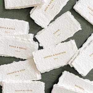 May include: A collection of handmade, textured white paper place cards with gold lettering. Each card displays names and table numbers, perfect for weddings or events. The cards are scattered on a green surface, creating an elegant and rustic aesthetic.