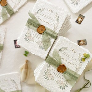 May include: A stack of white paper invitations with green and gold accents. The invitations feature a floral design with the text "Please join us as we celebrate the engagement and civil union of Carla Meli and Nico" and the date "Saturday, 29th April 2023".