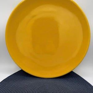 May include: A round, solid yellow plate. The plate is a bright, sunny yellow color. The plate is sitting on a dark blue, circular placemat. The plate is a simple, elegant design, suitable for serving food.