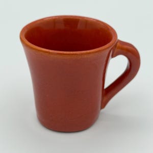 May include: A small, terracotta-colored ceramic mug with a curved handle. The mug has a slightly flared shape and a smooth, glossy finish. The interior of the mug is the same color as the exterior. The mug is designed for hot beverages.