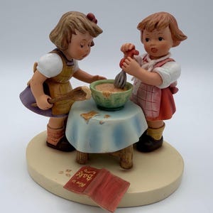 May include: A ceramic figurine depicting two young children baking. One child stirs a bowl of batter, while the other holds a wooden spoon. A small table with a blue tablecloth and a red cookbook with the words "How to Bake" are also present.