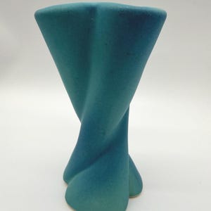 May include: A teal-colored, abstract ceramic vase with a twisted, hourglass-like design. The vase has a wide, square top that tapers down into a twisted body, and ends with a three-lobed base. The color gradient shifts from a darker teal at the top to a lighter shade at the base.