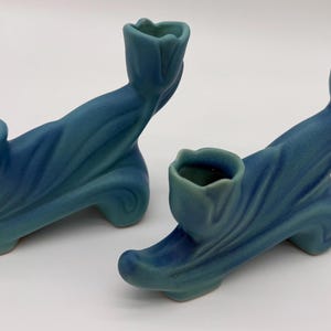May include: Two blue ceramic candle holders. Each has a curved base, a flowing, leaf-like design, and a tulip-shaped cup for a candle. The color is a blend of blue and teal.