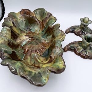 May include: A ceramic decorative set featuring a large, leaf-shaped bowl and two matching candle holders. The pieces are glazed in shades of green, brown, and yellow, with a glossy finish. The bowl has a scalloped edge, and the candle holders have a floral design.