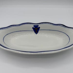 May include: White oval serving dish with a scalloped edge and a blue trim. A blue and gold emblem is centered on the rim. The dish is empty and sits on a white surface.