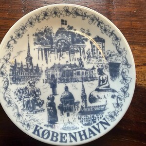 May include: A white ceramic plate with a blue illustration of Copenhagen landmarks, including a church spire, buildings, and the Little Mermaid statue. The plate is edged with a floral design and the word "KØBENHAVN" is printed at the bottom.