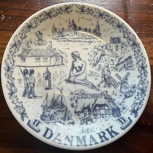 May include: A white ceramic plate with a blue illustration of Danish landmarks, including the Little Mermaid statue, a castle, and sailboats. The word "DENMARK" is printed at the bottom of the plate, with a floral border.